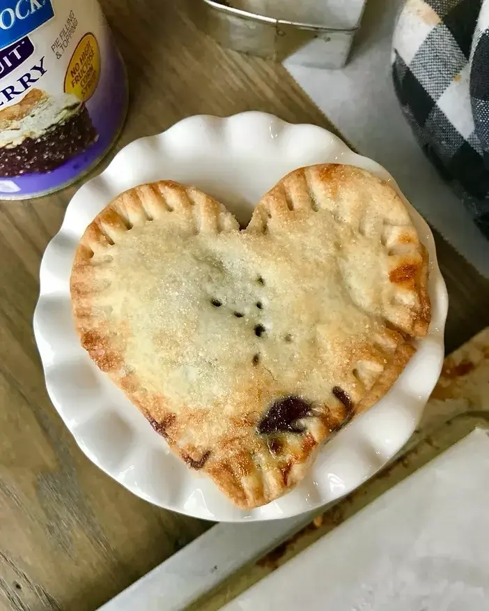 Heart-shaped pastry on a white plate, wooden table background. Nearby, a can reads "berry." Cozy kitchen vibe with a checkered cloth.