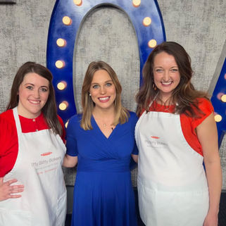 Three women in a kitchen, two wearing aprons. They are baking with dough on trays. The background features patterned tiles and shelves.