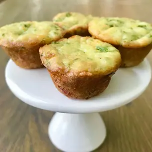 Four herb muffins on a white cake stand, set on a wooden table. The muffins are golden brown with green specks, creating a rustic vibe.
