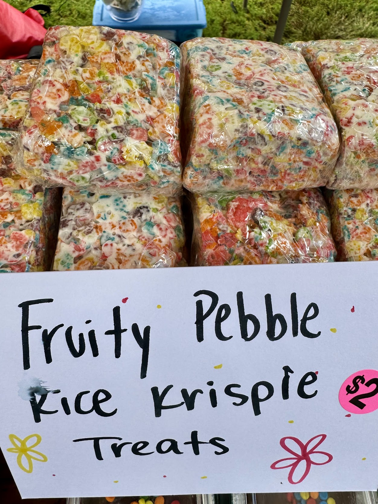 Colorful Fruity Pebble Rice Krispie treats in plastic wrap at an outdoor stall, with a hand-lettered $2 sign.