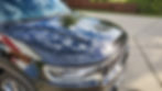 Shiny black car hood reflecting blue sky with clouds. "Wagoneer" text visible. Parked on concrete driveway with wooden fence in background.