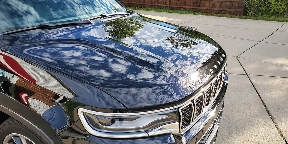 Shiny black car hood reflecting clouds and sky, with "Wagoneer" text visible. Parked in a driveway, wooden fence background. Calm mood.