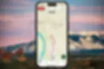 Phone with map app showing a hiking trail, in front of a scenic mountain background at sunset. Map displays distance and elevation.