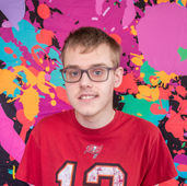 Headshot of student Ronan in front of a brightly colored splatter paint wall.