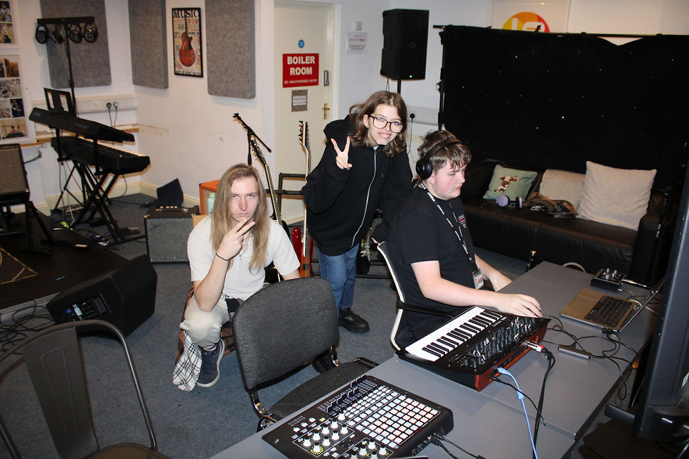 Three students in the SupaJam Canterbury music studio; one uses a keyboard, one flashes a peace sign, and another poses pensively. Instruments and "Boiler Room" sign visible.