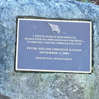 Photo of a memorial plaque mounted on a stone, inscribed with a dedication to Peter, Sue, and Christine Hanson, who lost their lives on September 11, 2001. The plaque features an engraved image of an American flag and the text, 'A tree planted in remembrance, shares with us a similar reason for being... to shelter, comfort, embrace and love.' Below, it reads, 'Groton will never forget.' The plaque is framed in a simple border and placed on a weathered stone surface.
