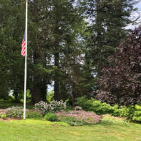 Photo of a peaceful garden area featuring a flower bed filled with pink and purple blooms, surrounding a white flagpole with an American flag flying at half-mast. The garden is nestled among tall, mature trees, with a mix of green foliage and dark-leaved trees creating a natural, serene backdrop. The well-maintained lawn in the foreground adds to the tranquil setting, making the garden a respectful and reflective space