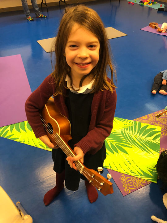Toddler stands on a mat holding a ukulele at a Little Groove ukulele class