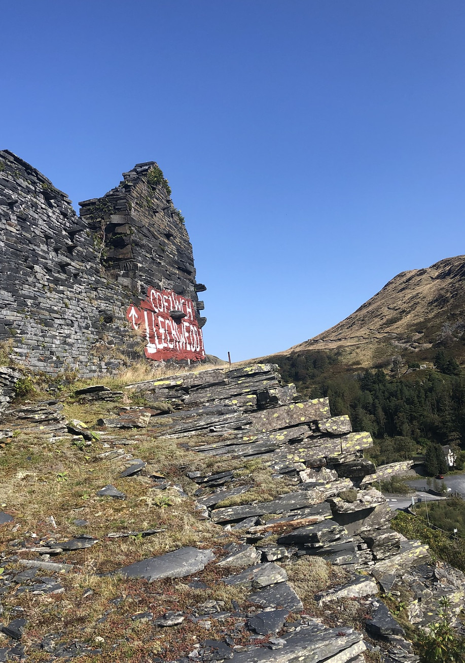 A photo of an old slate ruin with graffiti saying 'remember Llechwedd' against a blue sky,