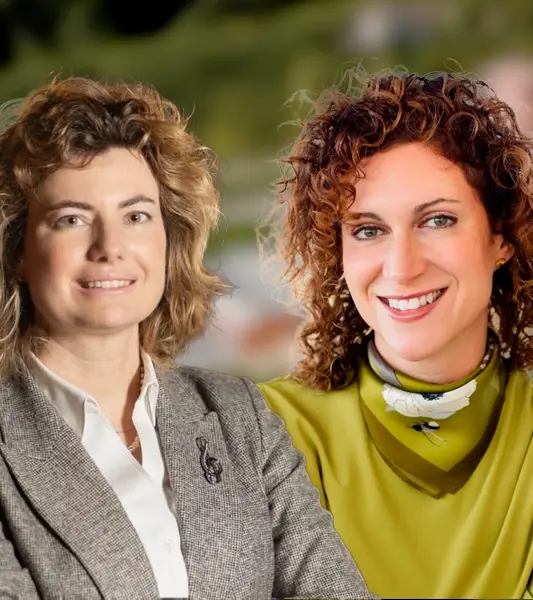 Two women smiling against a blurred outdoor background. The woman on the left is wearing a gray blazer over a white shirt, and the woman on the right is wearing a green blouse with a patterned scarf.