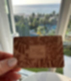 Hand holding a wooden hotel key card engraved with ‘The Palms Hotel & Spa’ and tropical leaf designs, with a window view of palm trees, a pool area, and the ocean in the background.