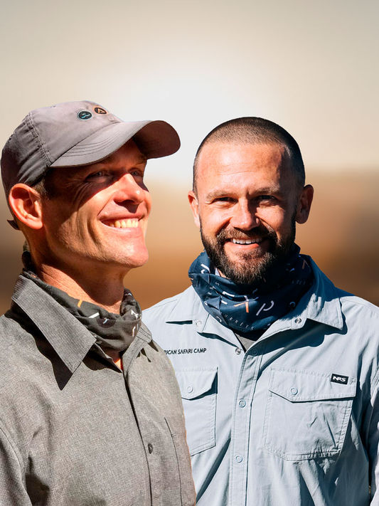 Two men smiling outdoors in a sunny, natural environment. The man on the left wears a gray cap, button-up shirt, and a patterned neck gaiter. The man on the right has a beard, short hair, and wears a light blue safari-style shirt with a dark patterned neck gaiter. The background features a blurred, warm-toned landscape suggesting a sunrise or sunset in a desert or savanna setting.