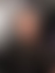 Portrait of a smiling woman with short curly hair wearing a black checkered blazer over a black top, standing in front of a historic interior staircase with ornate railings and a vintage atmosphere.