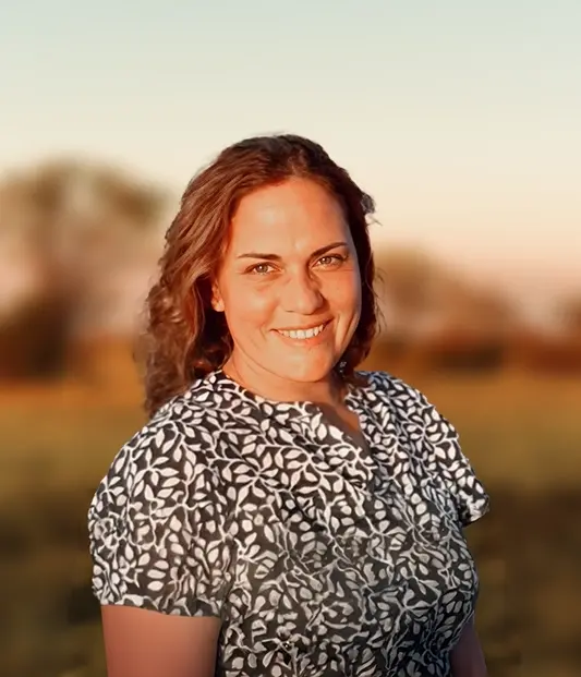 Smiling woman with wavy brown hair wearing a black-and-white patterned blouse, standing outdoors in a blurred field at sunset.