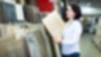 Woman in a store selects a light wood laminate. Shelves with assorted panels surround her. She's focused, wearing a white shirt.