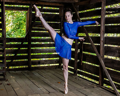Ballet dancer Emma en pointe in a blue costume inside a rustic tobacco barn.