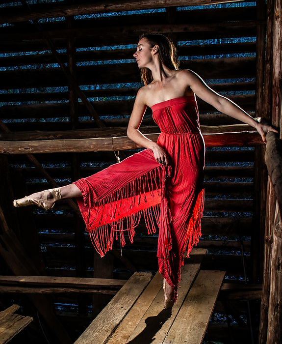 Ballet dancer Emma en pointe in a flowing red dress inside a rustic tobacco barn.