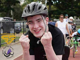 A boy wearing a helmet stands leaning on a running frame. He is facing the camera and smiling, with two raised fists in front of his face.