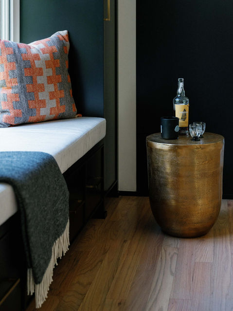 A cozy seating nook with dark green walls, a built-in bench, orange accent pillows, and a brass side table with a whiskey bottle and glassware