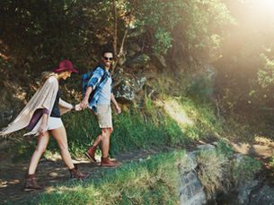 Man and woman hiking hand in hand on a sunny trail through lush greenery. She wears a red hat, both in casual attire, exuding joy and adventure.