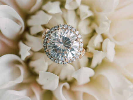 A round diamond ring serves as an engagement ring sitting on a neutral colored floral bloom