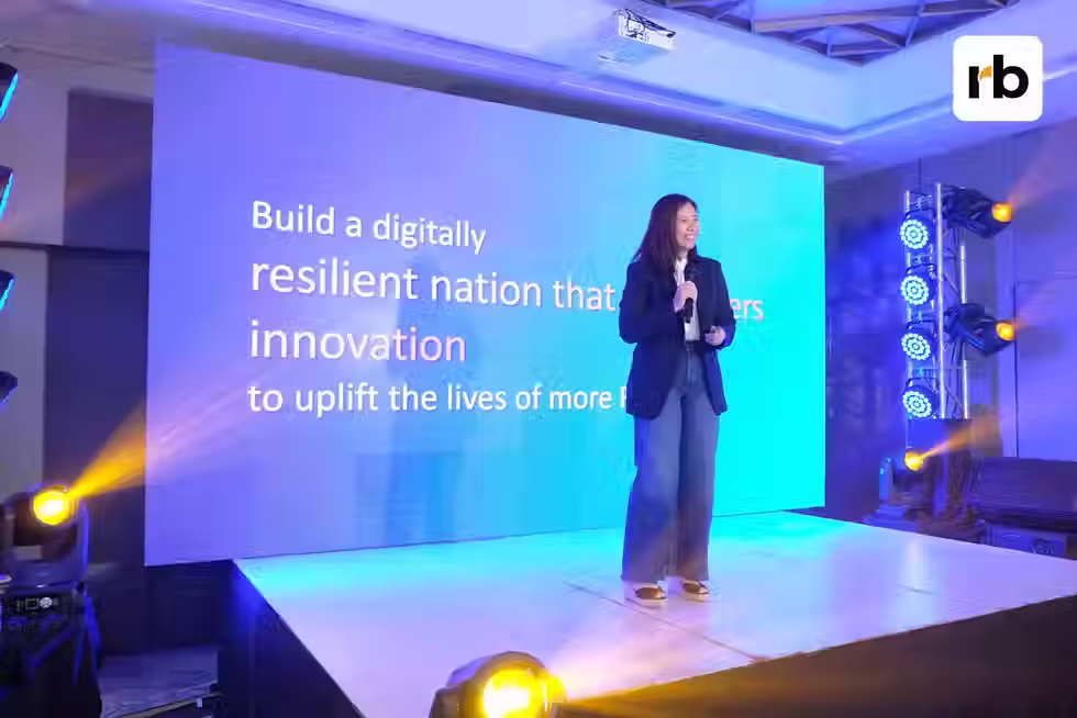 A woman speaks on stage with a microphone, standing in front of a large blue screen displaying text about digital resilience and innovation.