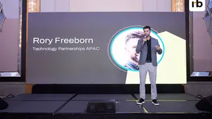 Man in gray suit speaks on stage. Behind him, a screen shows "Rory Freeborn, Technology Partnerships APAC." The mood is professional.