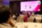 Panelists discuss customer experience on stage at a conference. Audience listens. Large screen displays event details. Room is dimly lit.