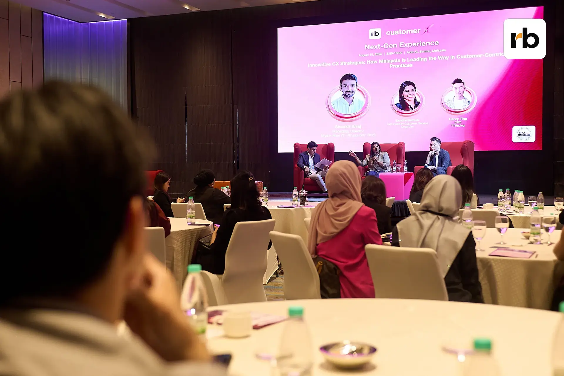  Attendees at tables participate in a customerX | rockbird media panel discussion, with a large screen displaying information in the background.