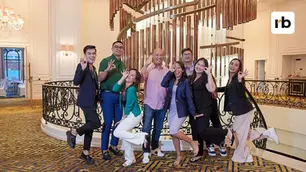 Eight people posing joyfully by a spiral staircase in an elegant room, all making "OK" gestures. The floor has a blue-gold pattern.