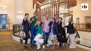 Eight people posing joyfully by a spiral staircase in an elegant room, all making "OK" gestures. The floor has a blue-gold pattern.