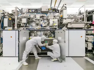 Technicians in white suits work on a complex machine in a sterile lab, examining components. Neutral tones, intricate equipment visible.