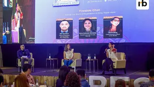 Panel discussion in a dimly lit room, four panelists speak on stage with a large screen behind them showing event details and portraits.