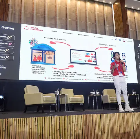 Person presenting on stage with large screen showing "Xchange Series" and "Wear Forward." Beige chairs and #GrowBeyond visible.