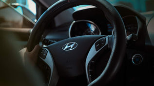 Close-up of a car steering wheel with a Hyundai logo, a hand gripping it. Buttons are visible. Interior is dimly lit with a calm atmosphere.