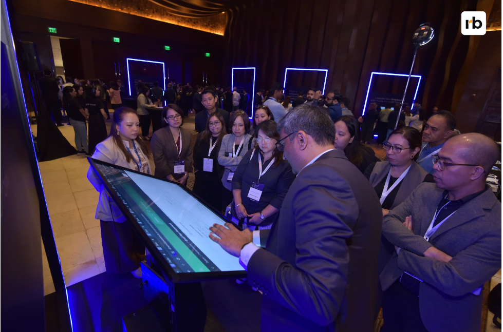 People gather around a large touchscreen in a dimly lit room, engaged and attentive. Blue neon frames illuminate the modern setting.