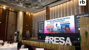A speaker on stage at a conference about offline and online retail in the Philippines. Large screen displays text and graphics. Bright, formal setting.