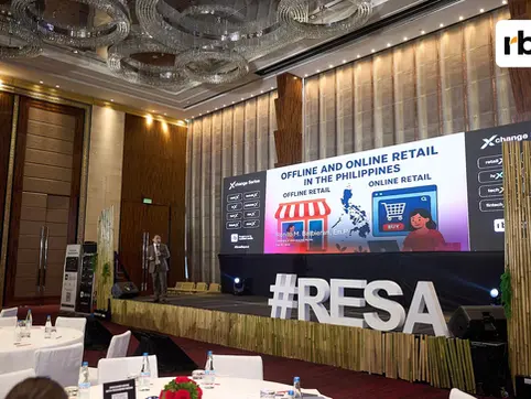 A speaker on stage at a conference about offline and online retail in the Philippines. Large screen displays text and graphics. Bright, formal setting.