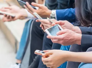 People sitting on steps, focused on their smartphones. Various colored clothing, hands holding phones. Busy, tech-focused atmosphere.