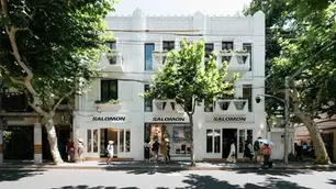 People walking by a white building with "Salomon" signs, shaded by large trees. Sunny day, relaxed outdoor atmosphere.