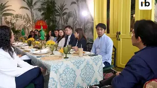 People sit around a table with green glasses, yellow flowers, and patterned cloth in a tropical-themed room. A yellow door is in the background.
