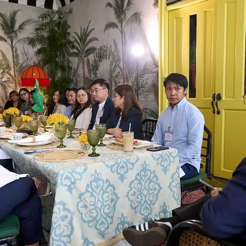 People sit around a table with green glasses, yellow flowers, and patterned cloth in a tropical-themed room. A yellow door is in the background.