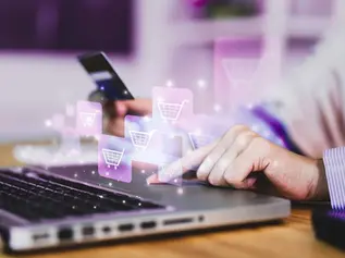 Person shopping online using a laptop and credit card. Digital shopping cart icons hover above the computer, suggesting e-commerce.