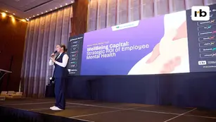 A woman speaks on stage with a backdrop displaying "WellBeing Capital: Strategic ROI of Employee Mental Health" in a modern conference hall.