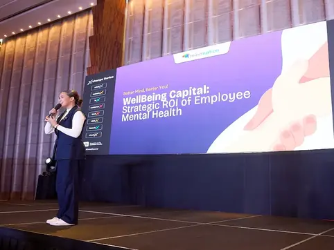 A woman speaks on stage with a backdrop displaying "WellBeing Capital: Strategic ROI of Employee Mental Health" in a modern conference hall.