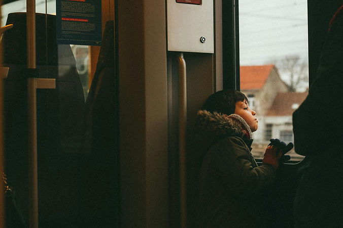 A young kid in a winter coat sits by a train window, holding a camera and looking out at blurred houses passing outside.