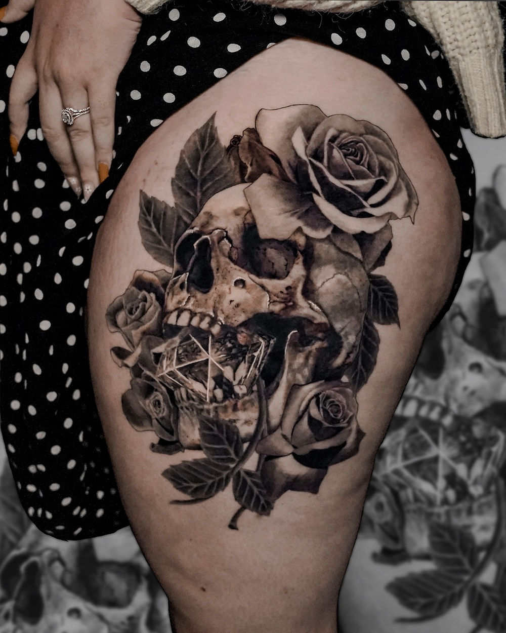 Large black and gray skull with a  diamond in its mouth surrounded by roses and leaves tattoo on woman's outer left thigh.