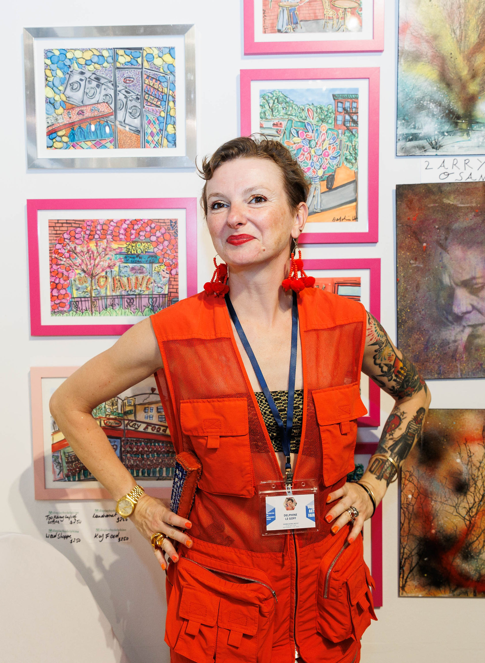 A woman in a red dress with tattoos stands confidently in front of framed colorful artworks.