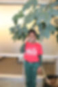 A little girl poses in red t-shirt in front of plant.