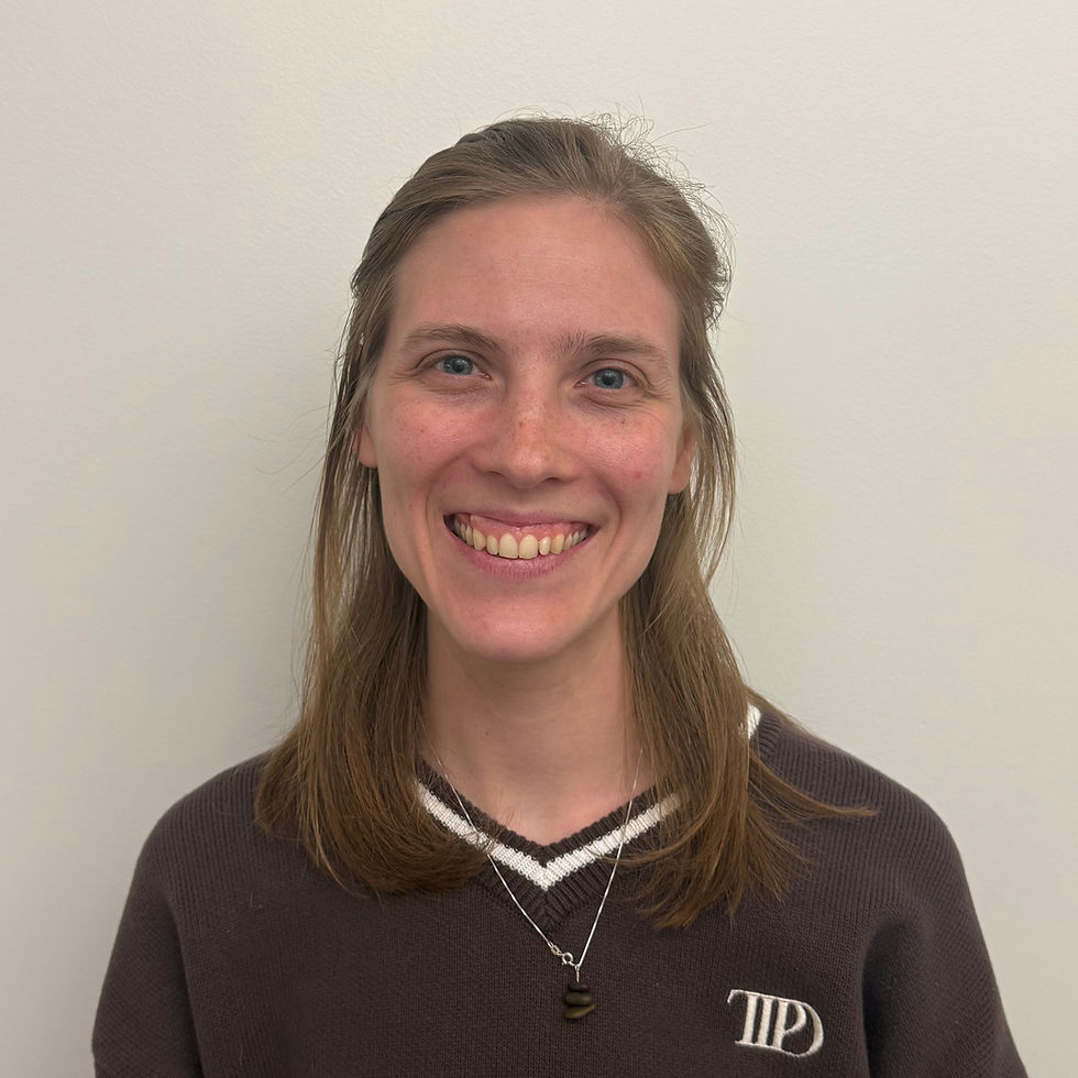 A fair-skinned American woman in her early 30s with blonde hair and blue eyes, smiling openly. She wears a dark brown V-neck sweater with a white trim and a necklace with a rock balancing pendant, standing in front of a white wall.